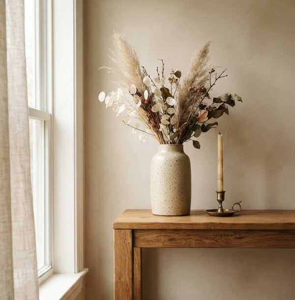 Oak console table by a window with a speckled stoneware vase of dried grasses and a brass candleholder.